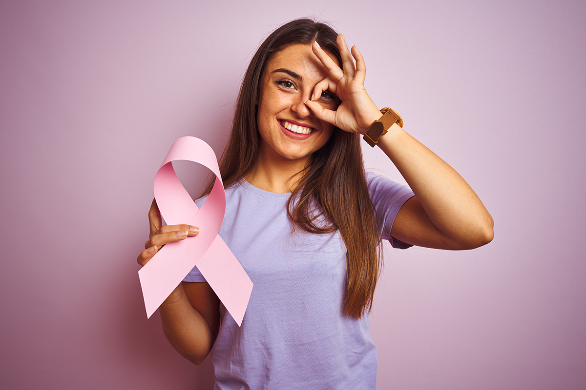 Beautiful woman holding cancer ribbon standing over isolated pink background with happy face smiling doing ok sign with hand on eye