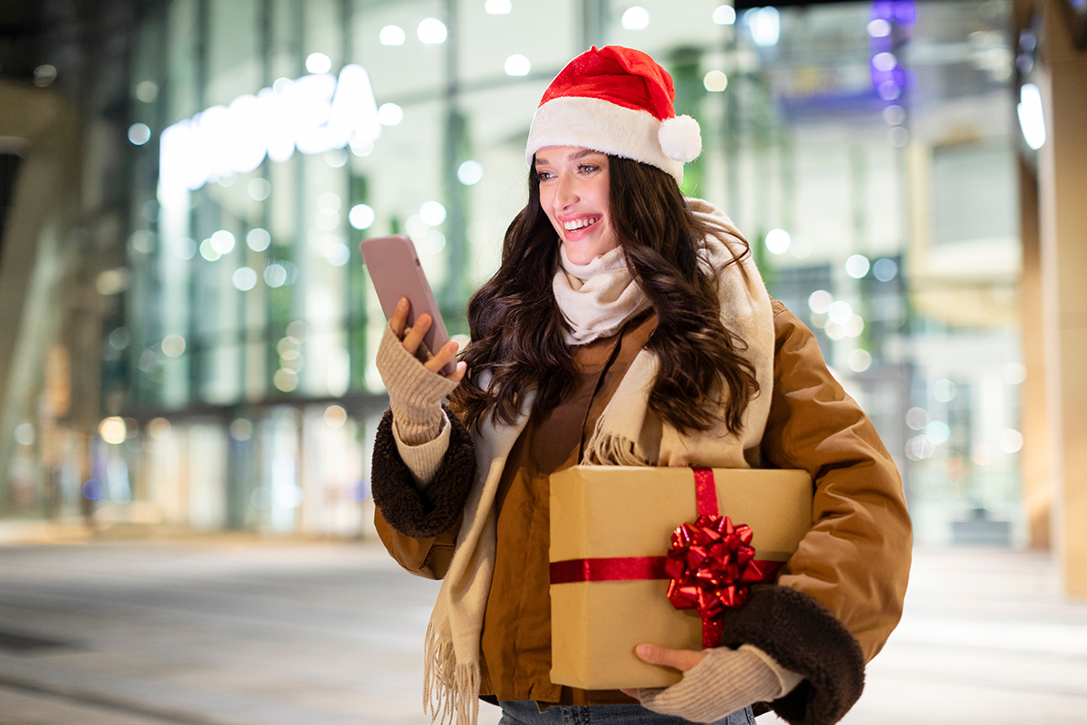 Joyful lady in Santa hat holding gift box and using smartphone while standing outdoors of shopping mall